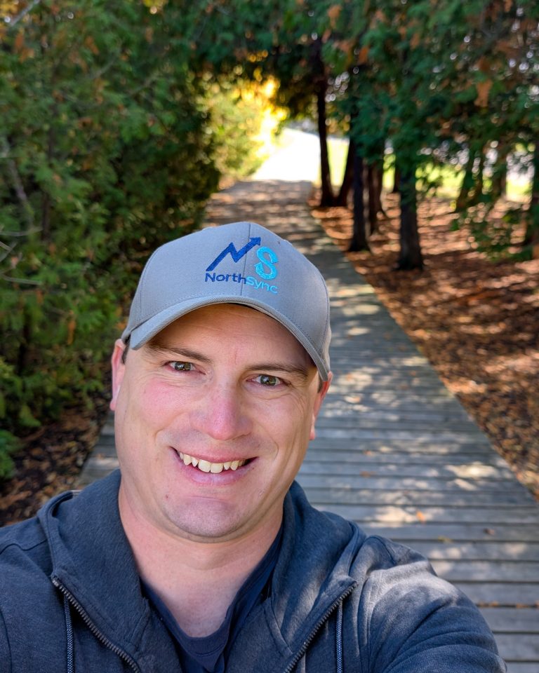 Home Mykal Petersen smiling on a forest boardwalk trail, wearing a NorthSync cap